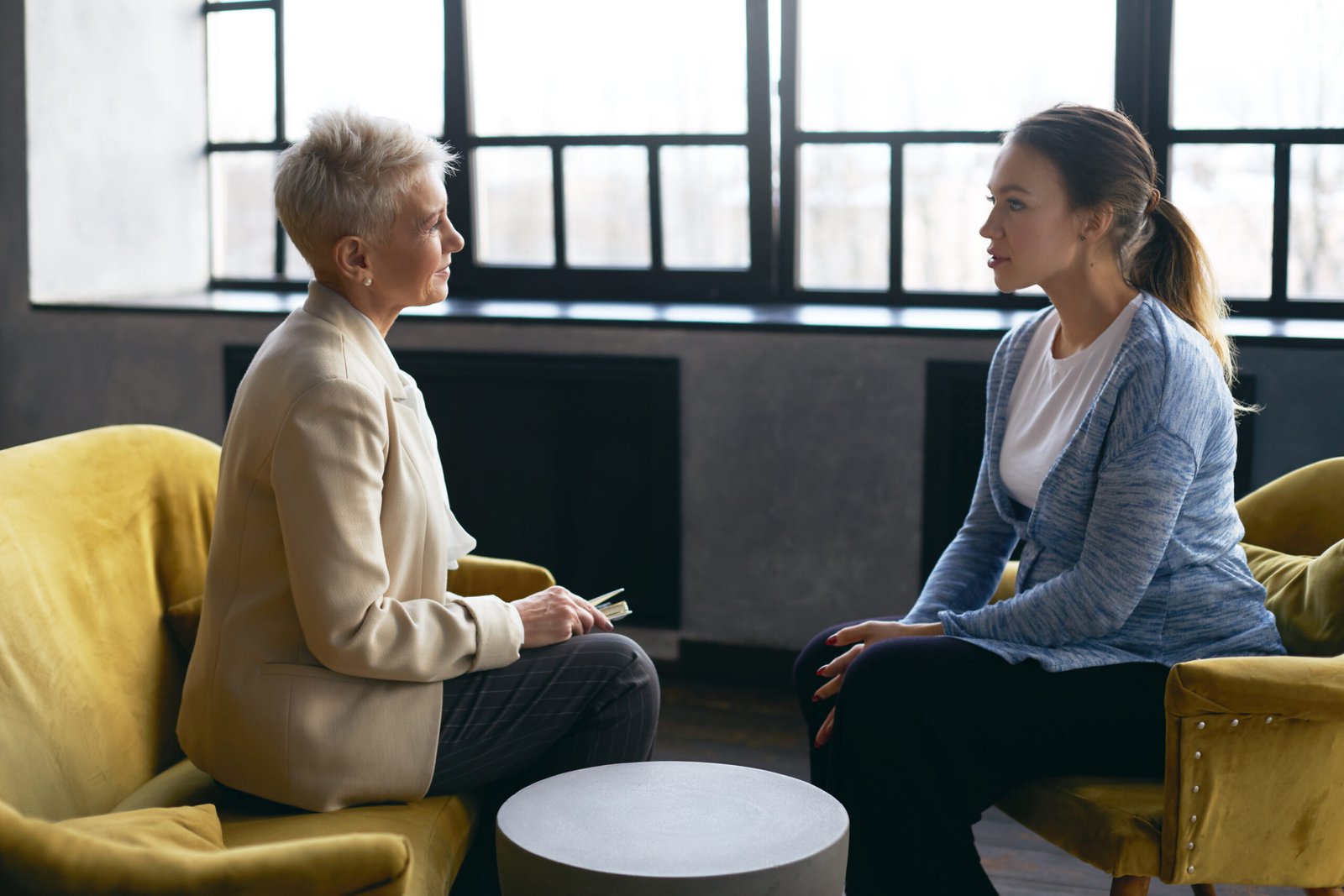 Side view image of middle aged female psychologist discusses the problems with young pregnant woman, sitting in armchairs, facing each other. Prenatal